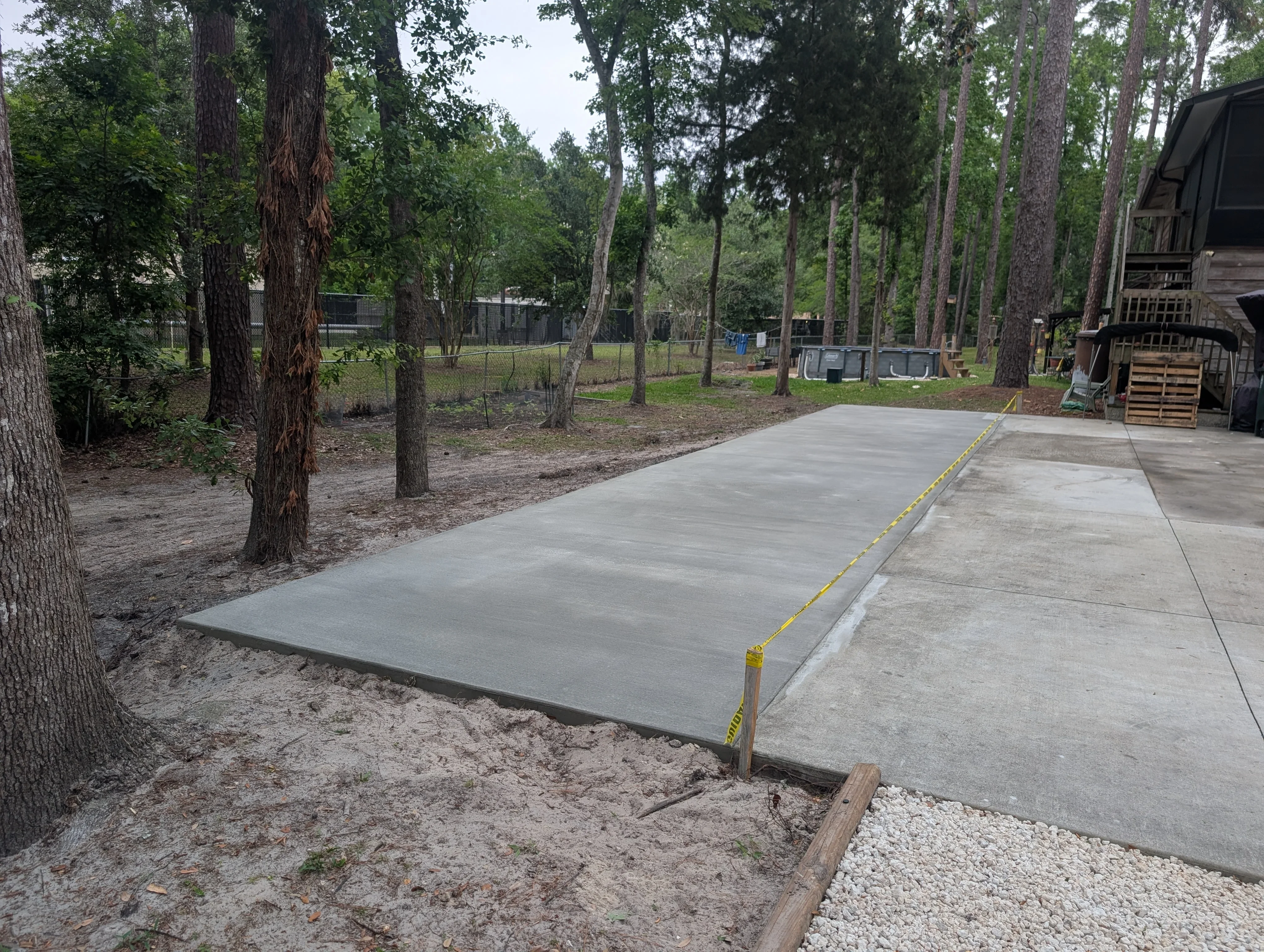Newly poured concrete pad with smooth gray finish, gravel base, and wooden forms in wooded area