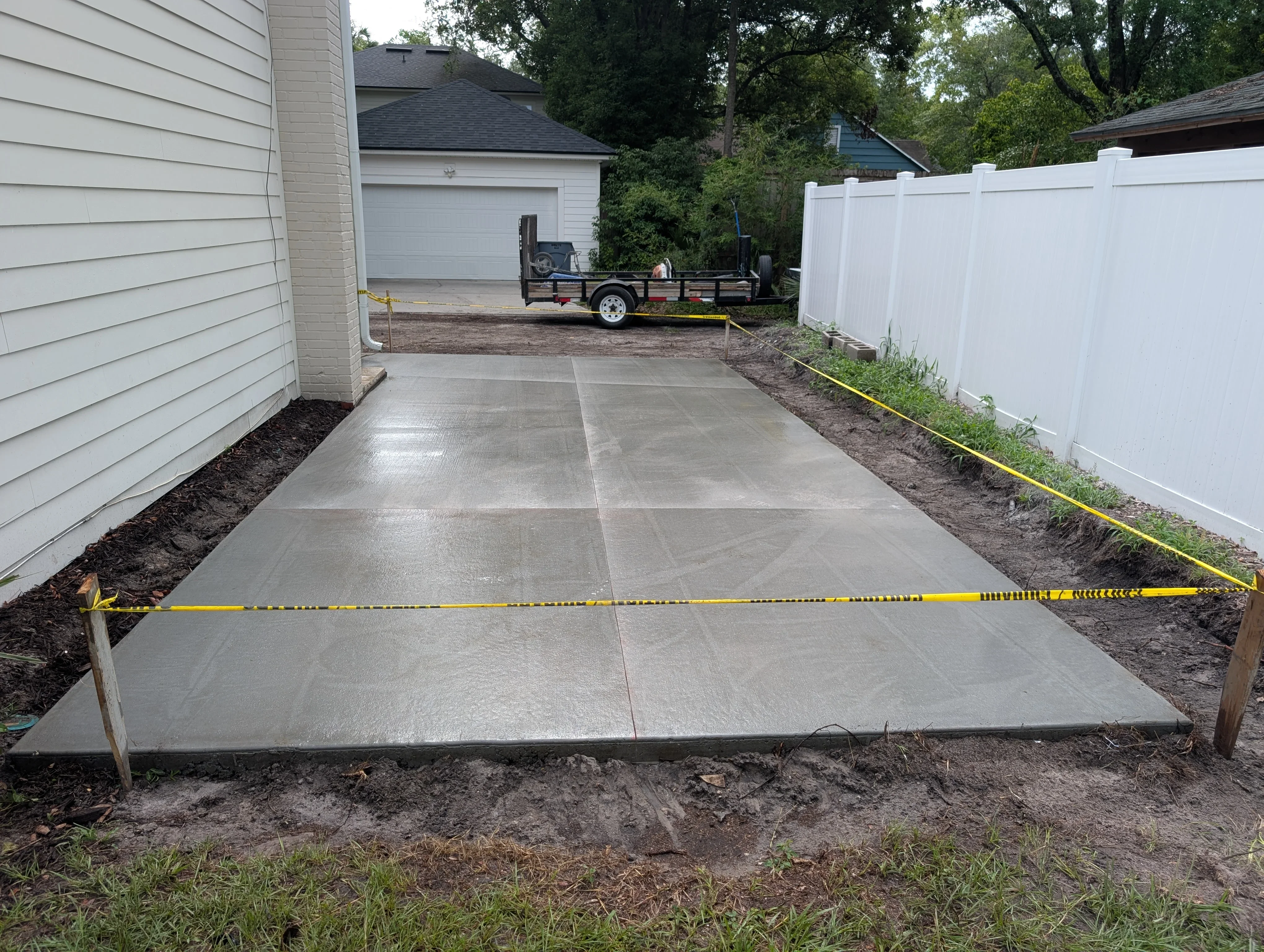 Gray concrete driveway with control joints, bordered by white fence and prepared soil beds