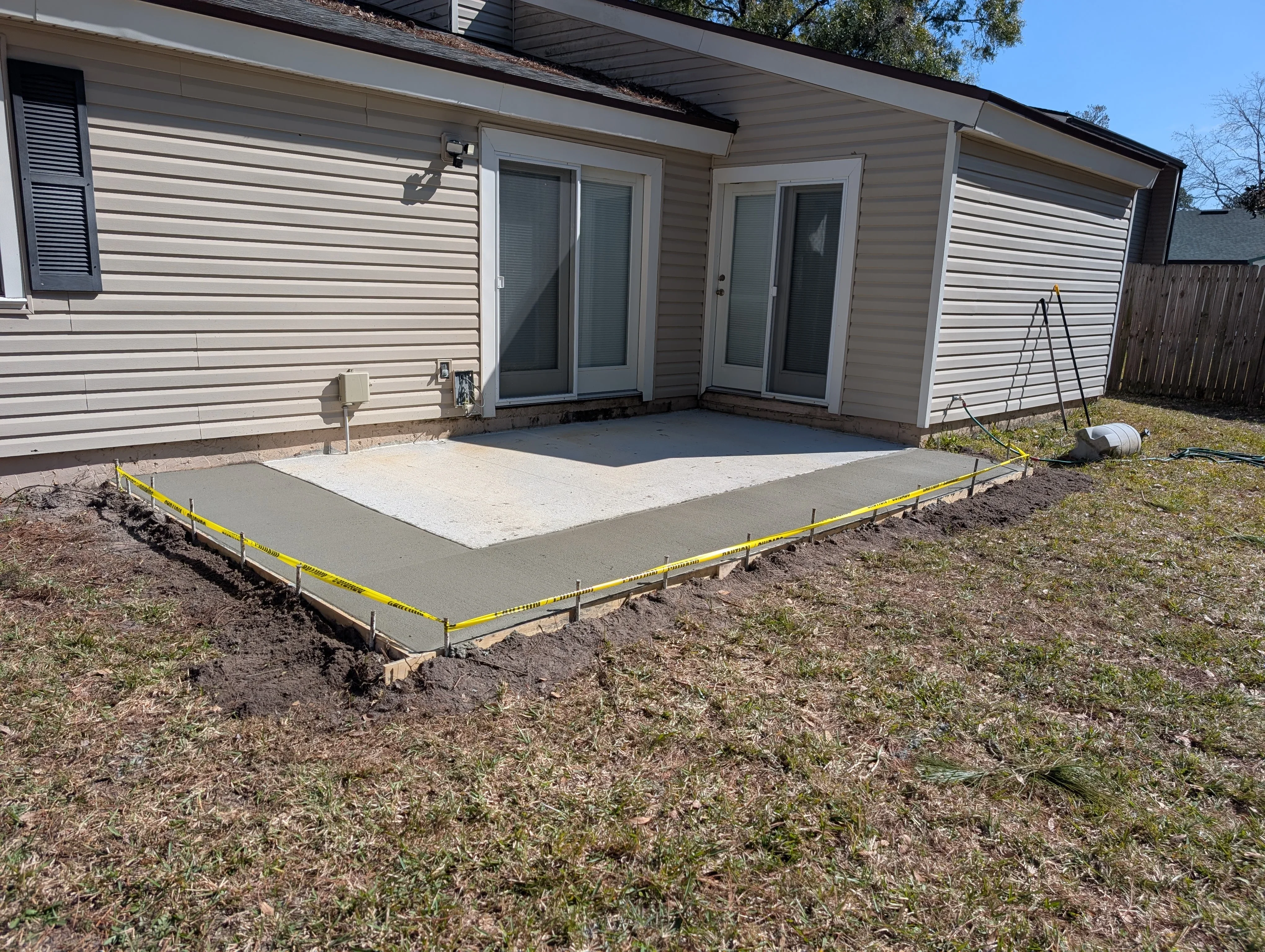 Newly poured concrete patio with light and dark concrete sections, yellow safety border, adjacent to house with sliding doors