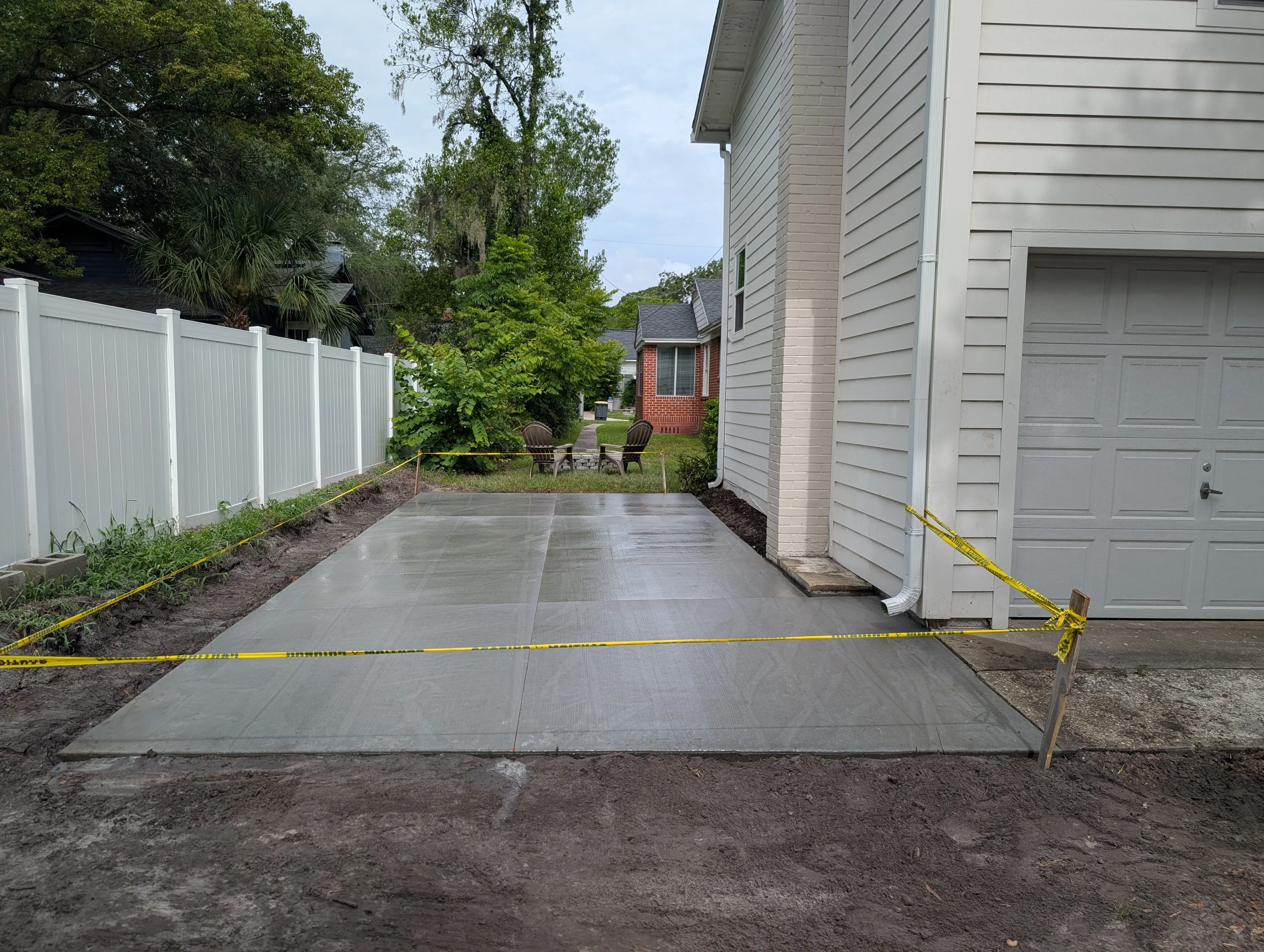 Newly poured concrete driveway with control joints, bordered by white vinyl fence and landscaped soil beds